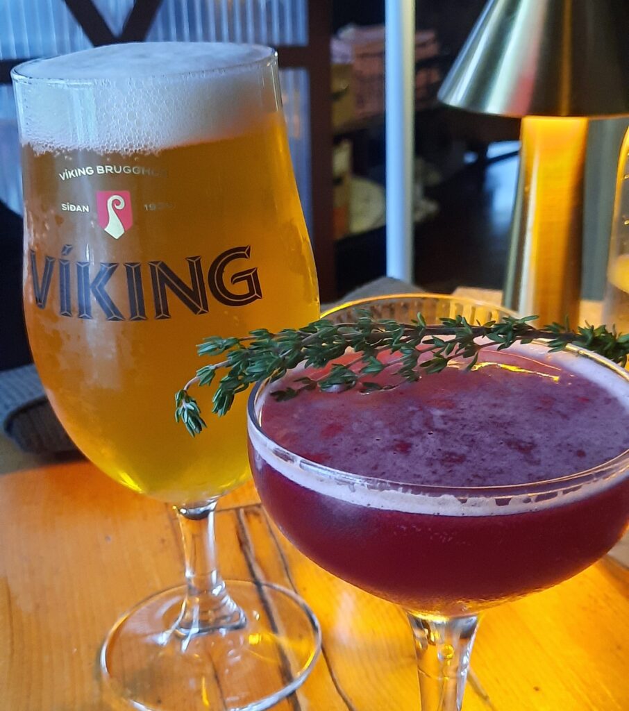 A purple crowberry gin with a thyme branch and behind it is a light coloured Viking beer. In the background is part of a lamp visible. All are on a wooden table.