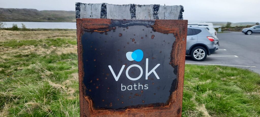 The sign of the Vök baths. It is sat on the parking lot so there is grass and cars behind it. 