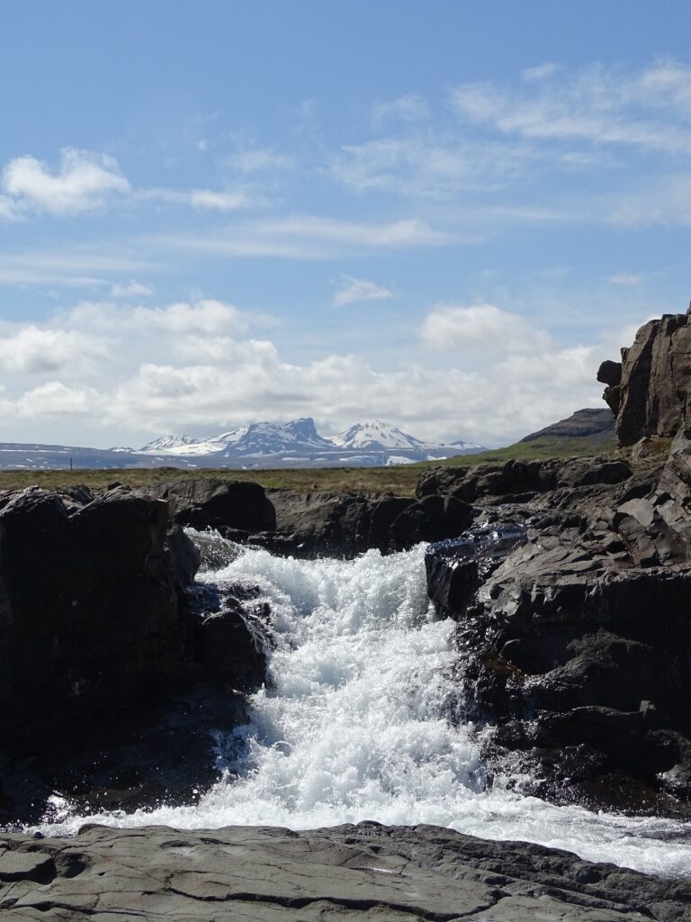 View at the foot of Lísafoss. The white water flows downs the dark coloured basalt rocks. In the background you see a glacier sticking out above the waterfall with blue skies and white clouds. 