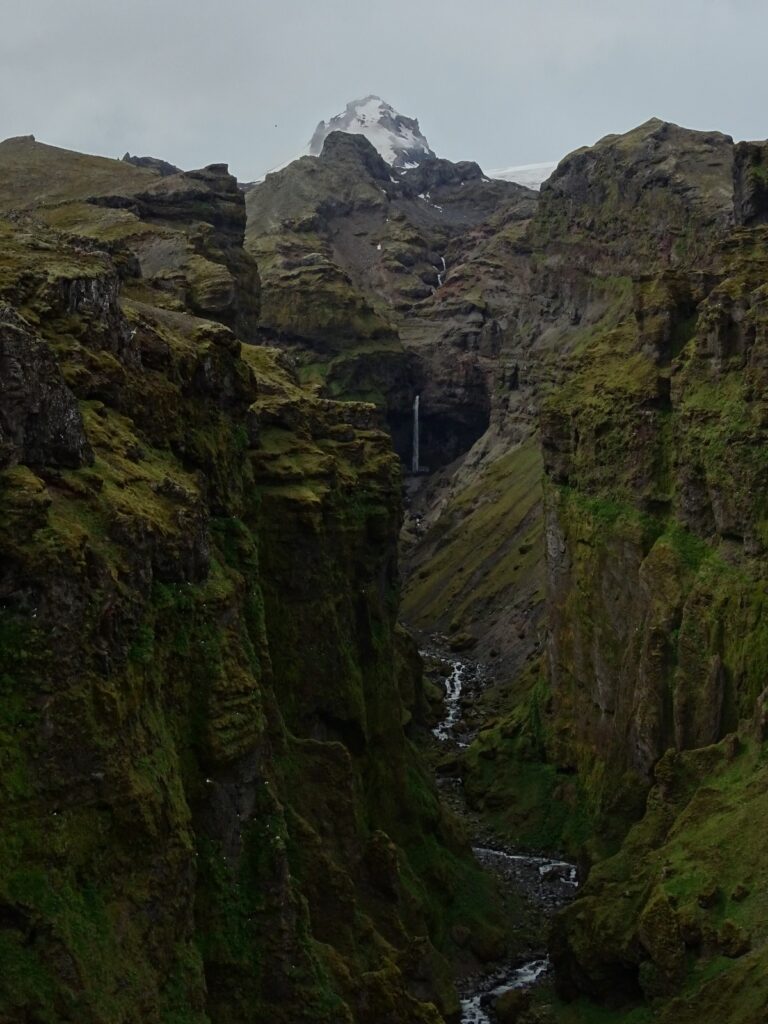 The picture shows the view into the Múlagljúfur Canyon. You can see the top of Rótarfjallshnúkur and the waterfall Hangandifoss underneath it . Green velvet moss covers the canyon walls and a snowy blanket covers the top of the mountain. 