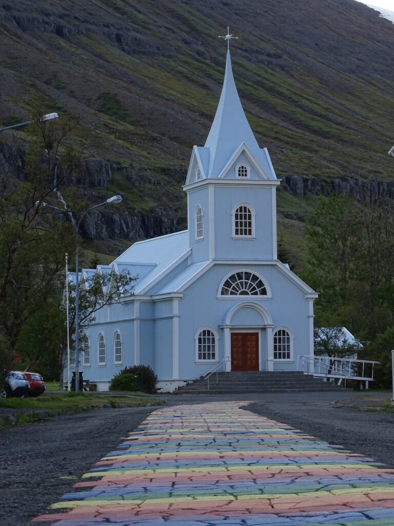 The light blue Seyðisfjarðarkirkja in Seyðisfjörður with the rainbowroad leading towards it. In the background you see the start of the Bjólfur Mountain.