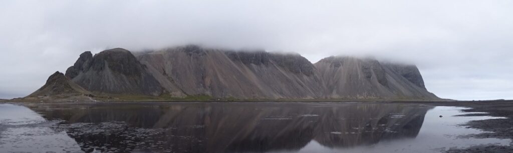 The tops of Vestrahorn Mountain are covered in a thick mist. With a thin layer of water remaining after low tide you can see the reflection of Vestrahorn.