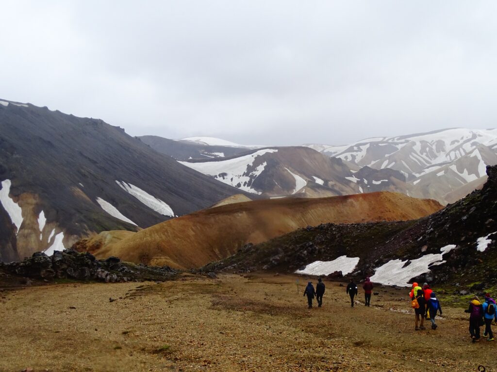 Walking trail in Landmannalaugar. Colourful mountains coloured in beige, orange, black, red and green covered with patches of white snow. A small group of hikers is walking on the right side of the picture. 