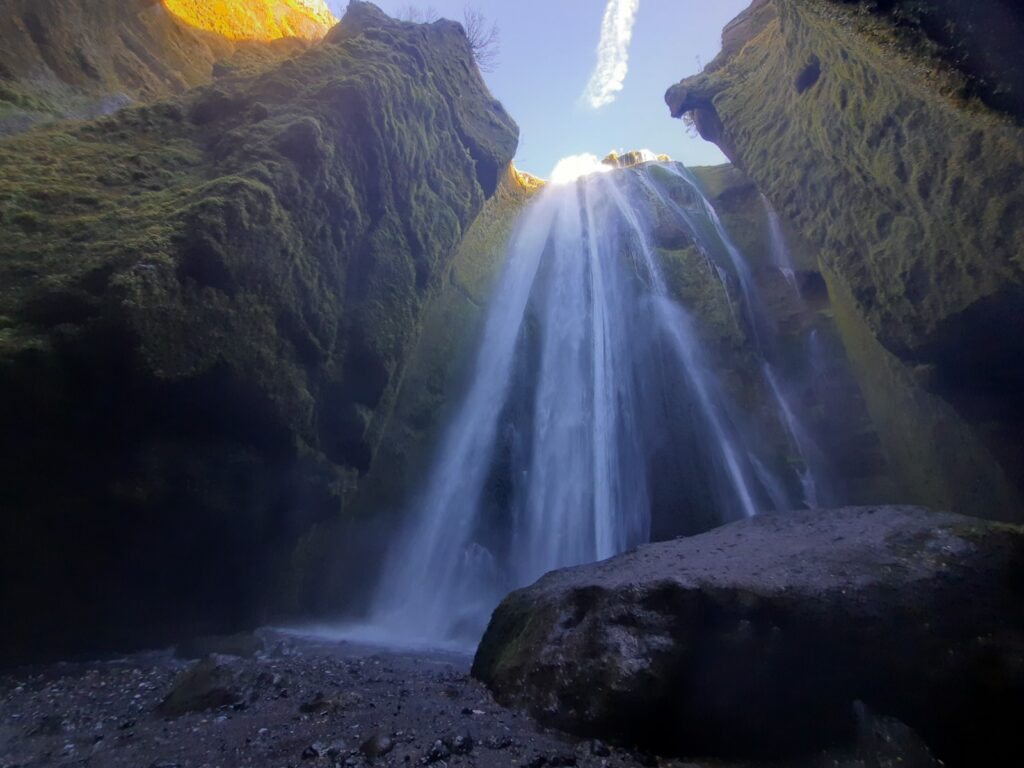 The waterfall of Gljufrabui falling down behind a big rock on the right side of the picture. through the narrow opening of the gorge you can see blue skies and a little bit of day light shines on the top of the left side of the gorge wall which is covered in soft green moss. 