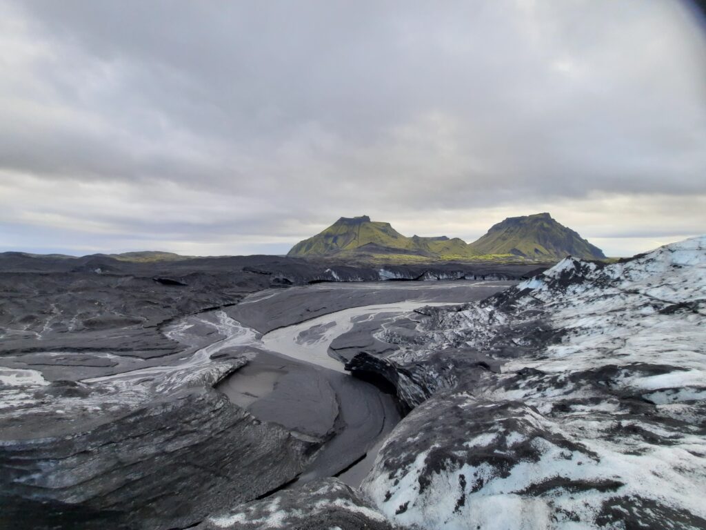 The black and white snow of the Katla glacier seems to flow out of the green hills in the back. The now greyish snow lays on the ashy black sand below with a small glacial stream going through it. The sky is covered in soft pastel coloured clouds. 