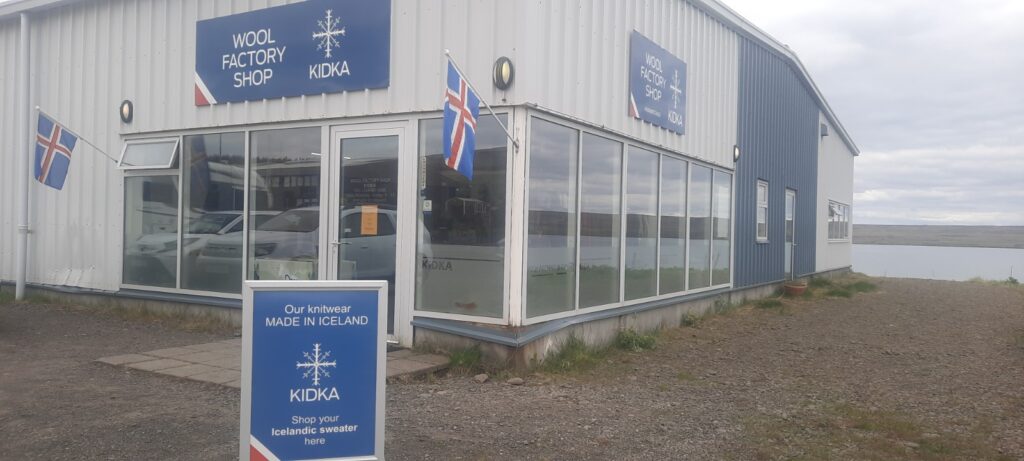 The front of the Kidka factory wool shop. Two Icelandic flag hang on its side. The building is white with blue boards holding the name. the bottom half of the building has big windows that allows you to look inside. Behind the building is water visible.