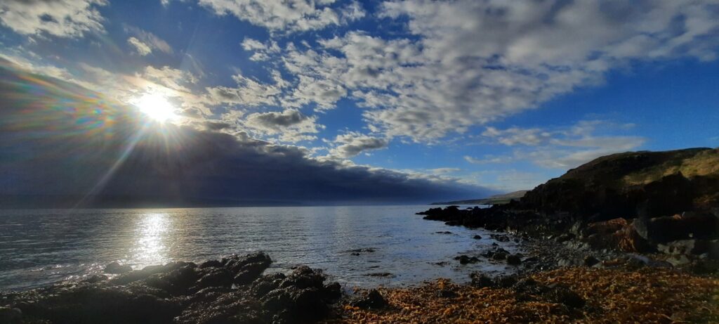 The picture shows a rocky beach covered in kelps. with behind it water. The sun is setting just over the top of the mountain ridge coming from the left. Giving the sun a last daylight shimmer. clouds float in a blue sky. 