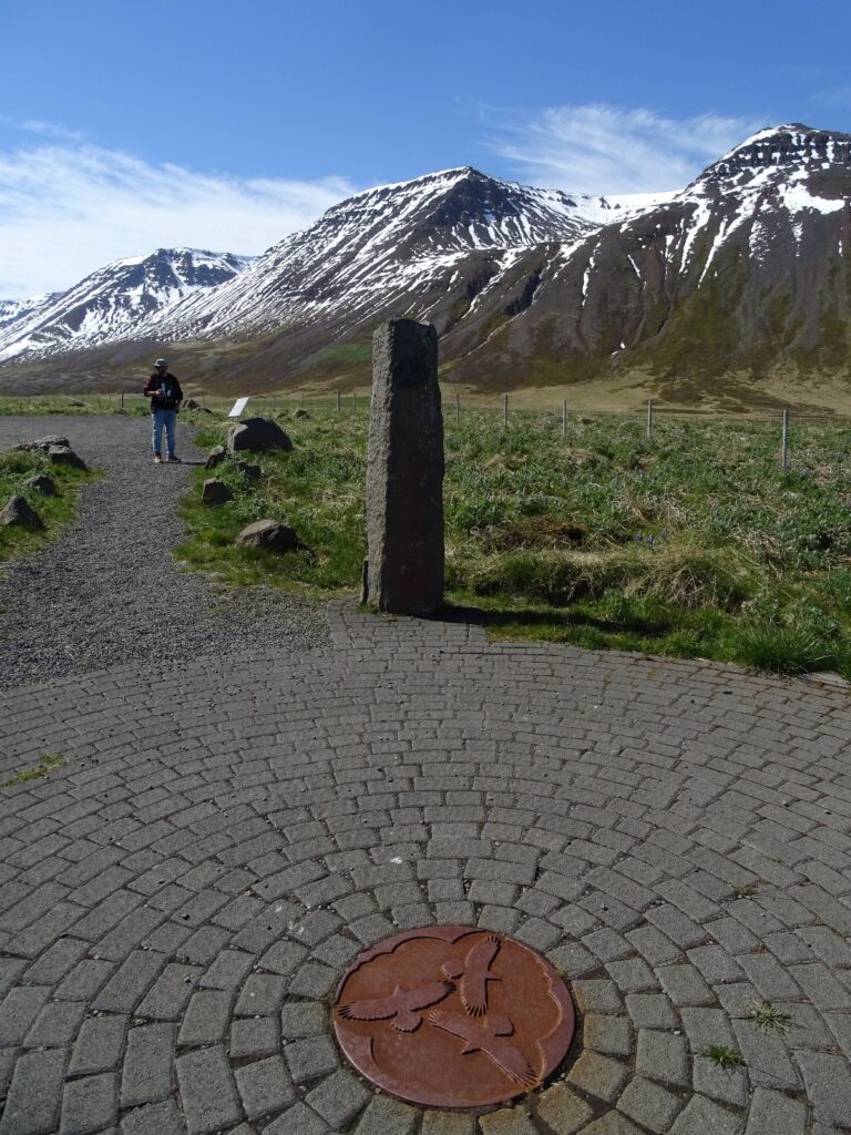 in the front in a round bronze looking plat in the middle of a round square. On this plate are 3 flying ravens. On the edge of the square, entrance point, is one pilar. There is a walking path on the left, with someone walking on it, going out of the picture. Around the square is grass land with not blooming lupins. All the way in the back are snow covered mountains and an almost clear blue sky. 