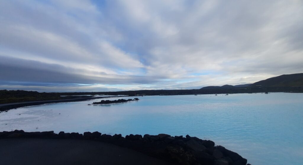 Blue water next to the blue lagoon seems even deeper blue than the sky above it. The pool is surrounded by dark coloured lava rocks. And on the edges the white silica is piled up. Clouds stretch over the blue and white pool. 