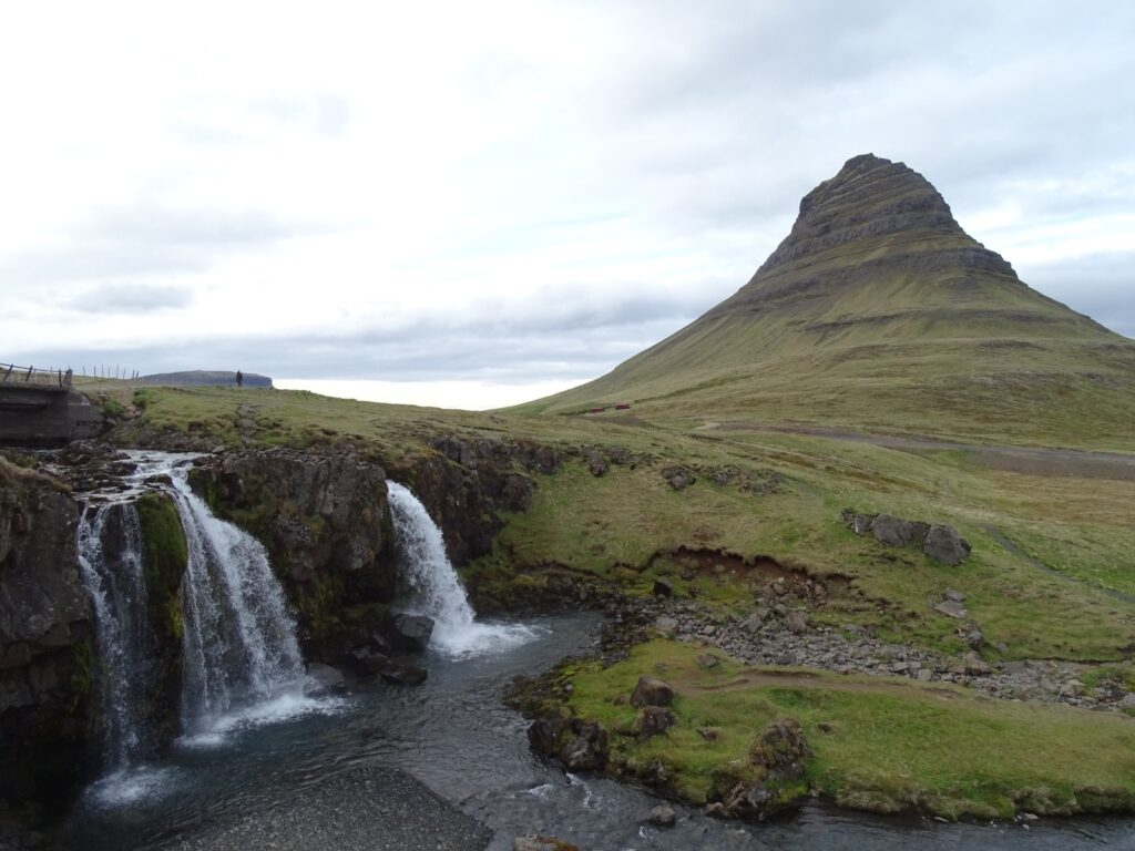 On the right is Mount Kirkjufell with on the left Kirkjufellsfoss falling into the pond. A cloud covered sky behind it all. 