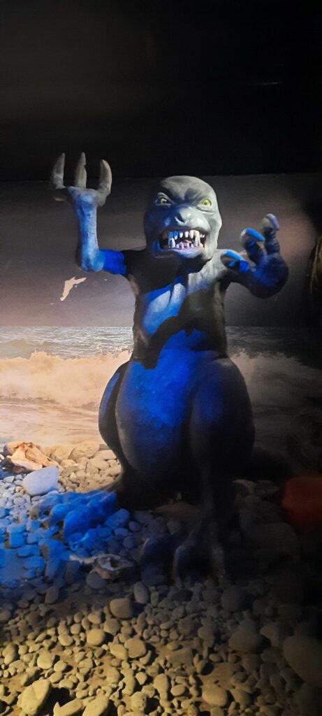 A life-size model of the Merman inside the museum. It is a grey monster with a bit of fur which makes the top look a bit like a werewolf, but without ears. It has thin arms with three claws. It has a pear-shaped bottom and big feet with 3 toes.