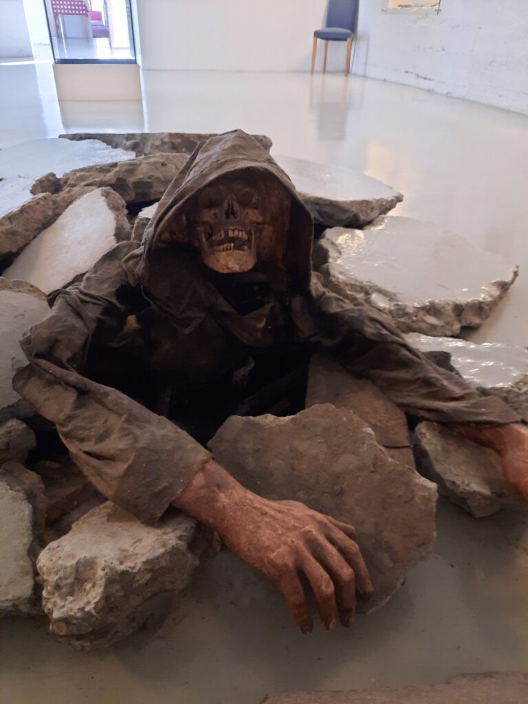 A skeleton in a dirty brown hooded cloak crawls out of the shiny white museum floor.