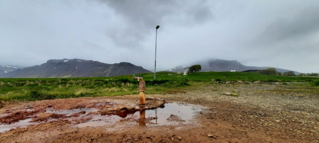 In the middle is the tap of the Ölkelduvatn Mineral Spring. The ground around it is orange stained from all the iron in the ground and in the water. Around it is some greenery and in the back are hills or mountains visible with a thin layer of mist over the tops. Above it is a grey, with clouds filled, sky.