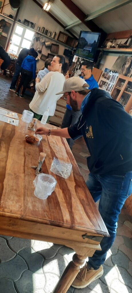 Someone is taking a piece of hákarl from the tasting table. The table is in the museum hall. The walls are filled with all kinds of things. In the upper right corner is a TV that shows pictures of the history. In the room are 5 other people looking around.  