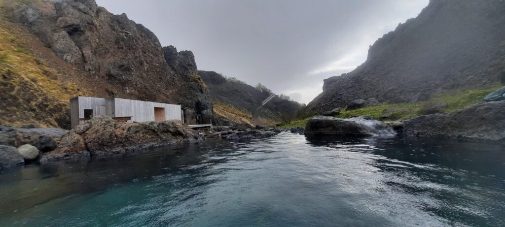 The view from the middle bath of the Húsafell Canyon baths. The water is in the front. On the left side is the little visible where your guide is during your stay. next to it is a darker building, this is the changing room. Everything is surrounded by the hills of the old volcanoes. 