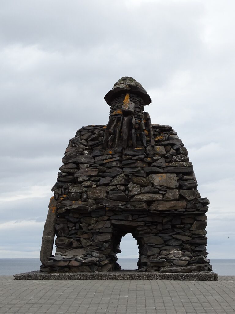 The stone statue of Bárður Snæfellsás. It looks like a thick arch with a head on top of it. The head has a big nose in the middle, with underneath it a beard. He is also wearing a hat. He is holding a plate in his right hand with is resting on the floor. It is standing on a square with in the back a little line of ocean  and a grey sky. 