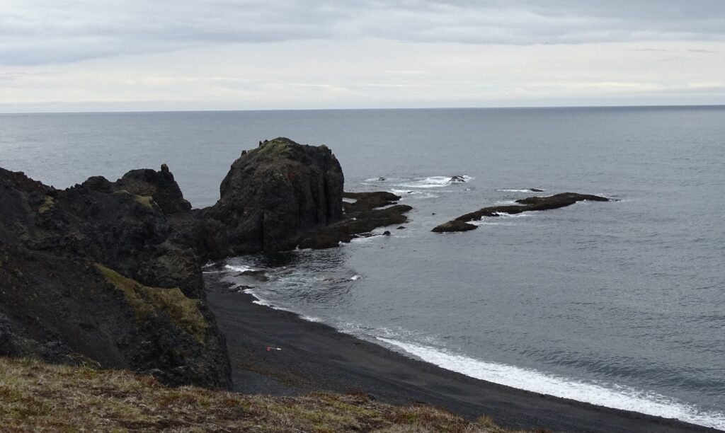 This is a top view of the left side of the Dritvík beach. There is a part of the dark beach visible, but the rocks look like sand. Going in to the sea is a rock formation, this is the Trolls' church. It is going into the grey ocean. In the front there is some greenery. 